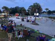 Families gather lawn chairs on grass and beach alike, to watch an outdoor movie.